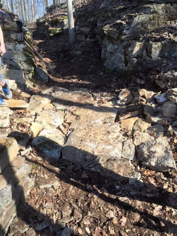 A rocky path leading into a wooded area, with scattered leaves on the ground and sunlight filtering through the trees. A shadow of a person is visible on the left side of the image, alongside a shovel leaning against a rock. O'bannon Woods mountain bike trail.