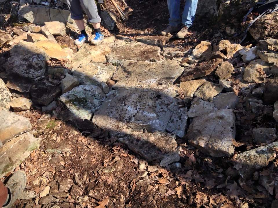 A partially cleared pathway made of large stones, surrounded by smaller rocks and scattered autumn leaves. Two people are visible, standing on the path, with one wearing shorts and athletic shoes. The setting appears to be a wooded area with a natural, earthy background. O'bannon Woods mountain bike trail.
