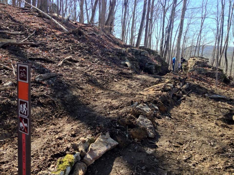 A dirt trail in a wooded area, flanked by trees with bare branches. A signpost indicating trail rules for biking and hiking is visible in the foreground. Two people are seen in the background, navigating the trail among rocky terrain and scattered leaves. O'bannon Woods mountain bike trail.