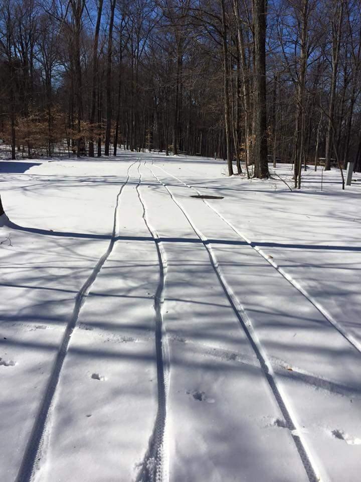 A snowy path through a forest, showing multiple tire tracks in the fresh snow leading into the trees under a clear blue sky. Shadows from the bare trees create a patterned effect on the snow. O'bannon Woods mountain bike trail.