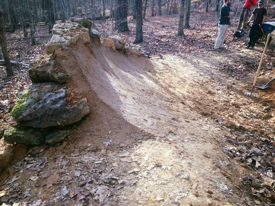 A dirt jump ramp constructed with a rock base, surrounded by autumn foliage and trees in a wooded area. In the background, individuals are seen engaging in outdoor activities, possibly preparing the jump or enjoying the natural setting. O'bannon Woods mountain bike trail.