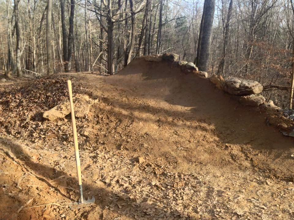 A cleared dirt path in a wooded area, featuring an elevated section built up with rocks. A digging tool is inserted into the ground nearby, surrounded by a layer of fallen leaves and bare trees. The setting suggests ongoing construction or trail maintenance. O'bannon Woods mountain bike trail.