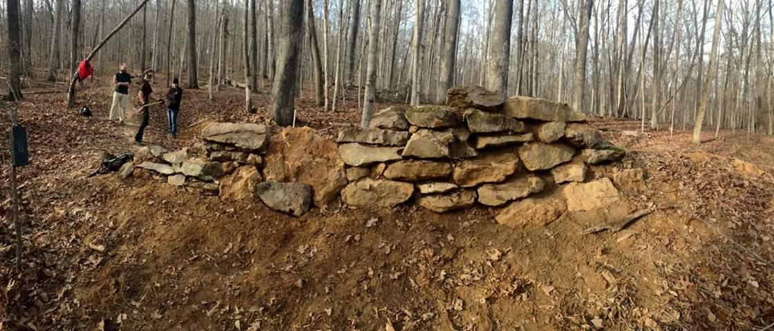A rocky wall constructed from large stones, situated in a wooded area with bare trees and fallen leaves. Three people stand nearby, engaged in outdoor activity. O'bannon Woods mountain bike trail.