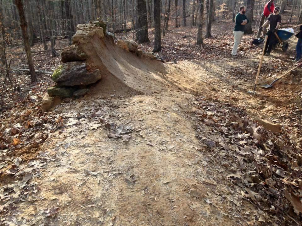 A dirt ramp with a rock formation at the top surrounded by trees in a wooded area. Several people are gathered nearby, some with tools, as they work on the trail or ramp. The ground is covered with fallen leaves. O'bannon Woods mountain bike trail.