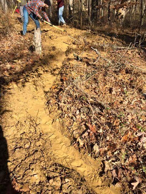 Two people work on constructing or maintaining a dirt trail in a wooded area. The trail is marked by freshly disturbed soil and scattered fallen leaves, surrounded by trees and underbrush. One person is using a tool to shape the trail, while the other is positioned slightly behind. The sunlight filters through the trees, casting shadows on the ground. O'bannon Woods mountain bike trail.