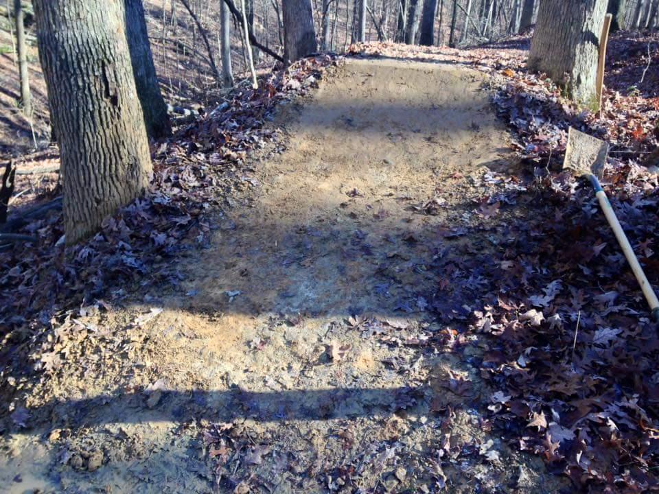 A dirt path surrounded by trees in a wooded area, covered with fallen leaves. A shovel is resting on the ground beside the path. O'bannon Woods mountain bike trail.