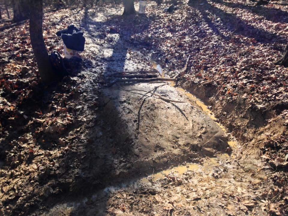 A muddy patch in a forested area with fallen leaves scattered on the ground, showing a small water puddle. A few sticks are placed across the muddy surface, and a backpack can be seen in the background near a tree. O'bannon Woods mountain bike trail.