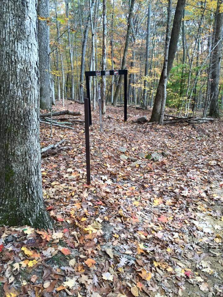 A wooden swing set with a black frame stands among colorful autumn leaves in a wooded area. The surrounding trees have yellow and green foliage, creating a serene natural setting. The ground is covered with fallen leaves, indicating the season. O'bannon Woods mountain bike trail.