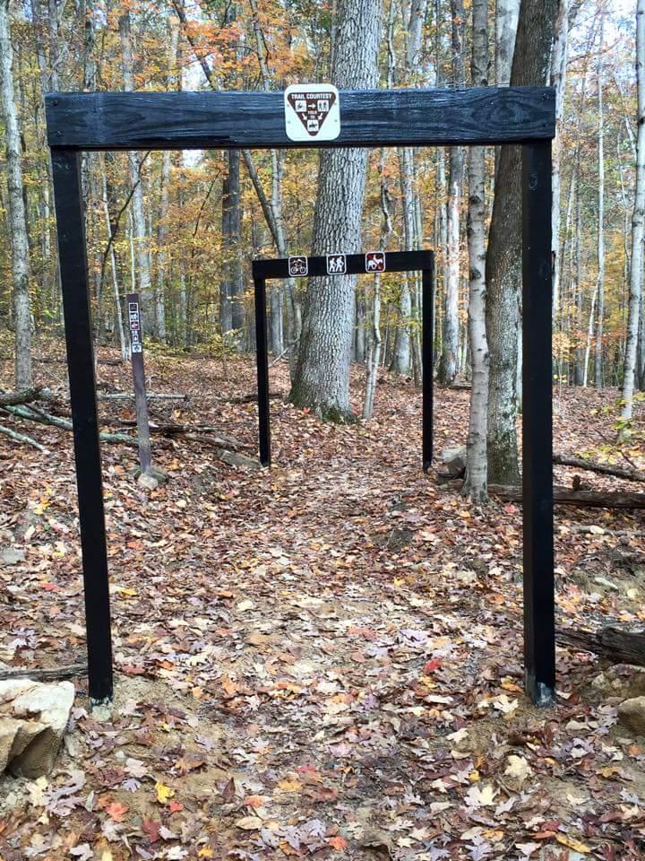 A trail entrance marked by wooden arches in a forest, surrounded by vibrant autumn foliage and fallen leaves. The arches have signs indicating trail etiquette, including symbols for biking, hiking, and restricted activities. The path leads deeper into the wooded area. O'bannon Woods mountain bike trail.