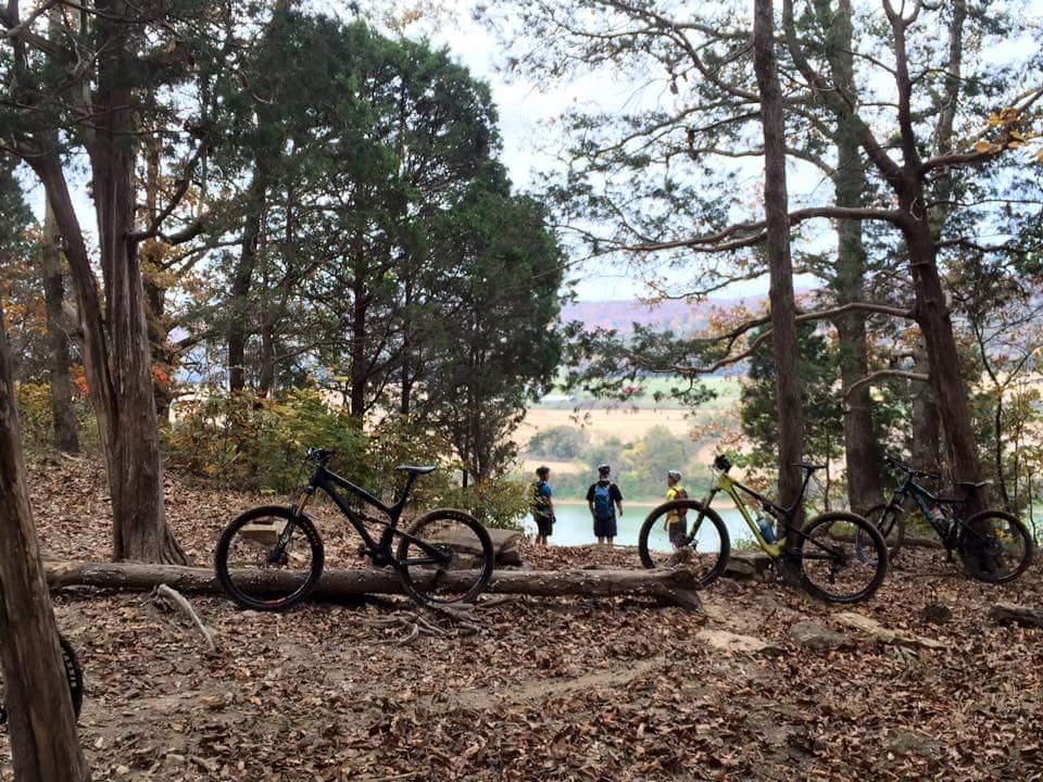 A scenic view of a forested area with two mountain bikes leaning against a log in the foreground. In the background, two people are standing near a body of water, surrounded by tall trees and autumn foliage. The atmosphere is peaceful, showcasing nature's beauty. O'bannon Woods mountain bike trail.