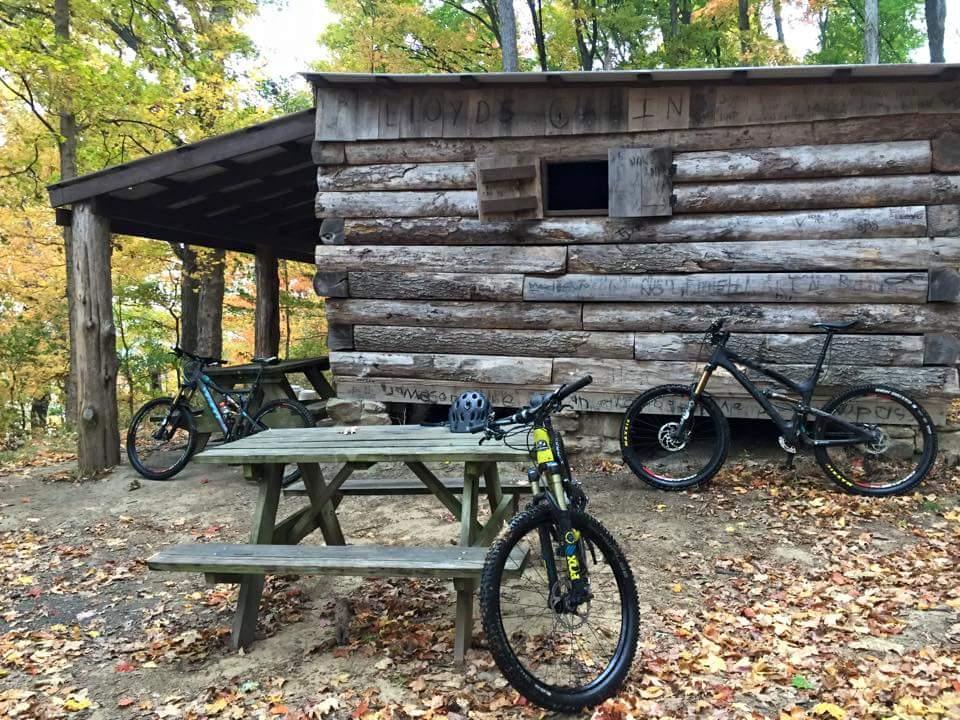 A rustic log cabin surrounded by colorful autumn foliage, featuring a wooden picnic table in the foreground. Two mountain bikes are parked nearby, one with a black frame and the other with a mix of blue and green. A helmet rests on the picnic table, adding to the outdoor adventure scene. O'bannon Woods mountain bike trail.