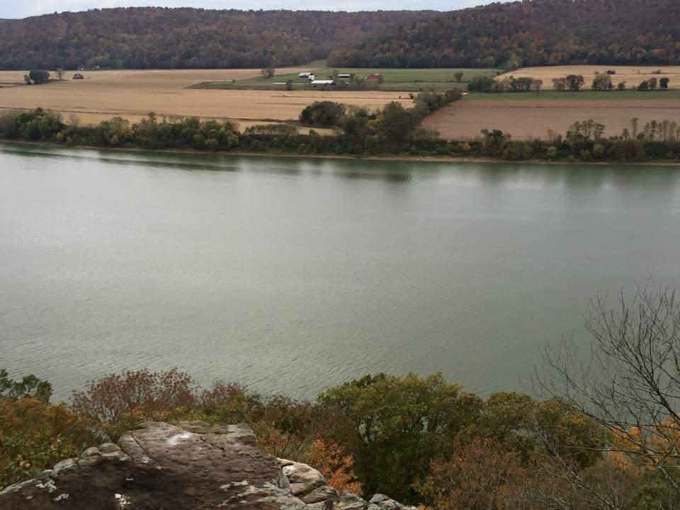 A scenic view of a river bordered by farmland and autumn foliage. The water reflects the soft gray sky, while rolling hills and trees in various shades of green and gold surround the landscape. In the distance, small farm structures can be seen against the backdrop of fields. O'bannon Woods mountain bike trail.