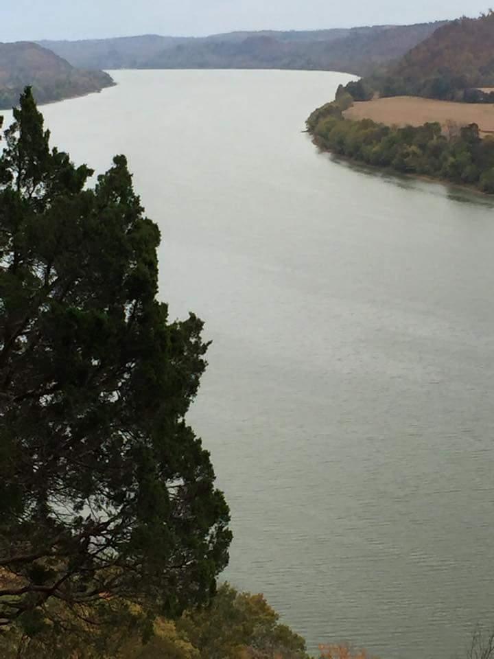 A serene view of a winding river surrounded by hilly terrain and lush greenery, with a prominent evergreen tree in the foreground. The landscape exhibits calm water reflecting the soft light, creating a peaceful natural setting. O'bannon Woods mountain bike trail.