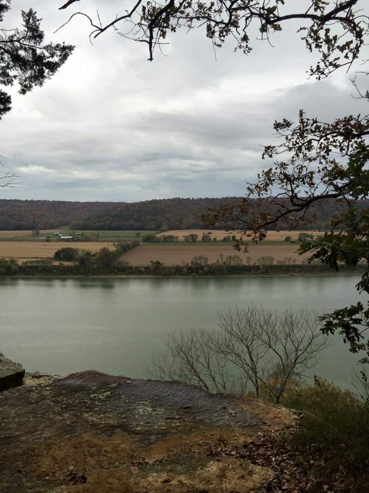 A scenic view of a river bordered by fields and distant hills under a cloudy sky. The foreground features rocky terrain with sparse trees, while the river reflects the overcast sky. The landscape displays autumnal colors in the fields and foliage. O'bannon Woods mountain bike trail.
