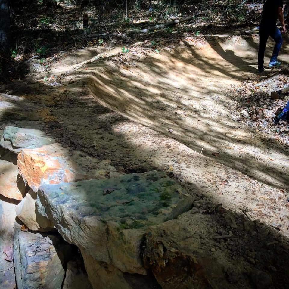 A sunlit dirt trail in a wooded area, with uneven, sloped edges and scattered fallen leaves. A rocky ledge runs along one side of the trail, and a person can be seen walking in the background. Shadows from nearby trees create a dappled light effect on the ground. O'bannon Woods mountain bike trail.