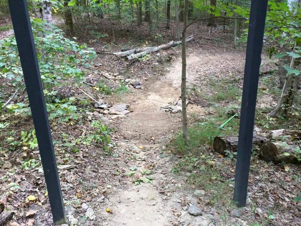 A dirt path winding through a wooded area, framed by two vertical posts. The ground is covered with leaves and small rocks, leading into a natural trail surrounded by greenery and tree trunks. O'bannon Woods mountain bike trail.