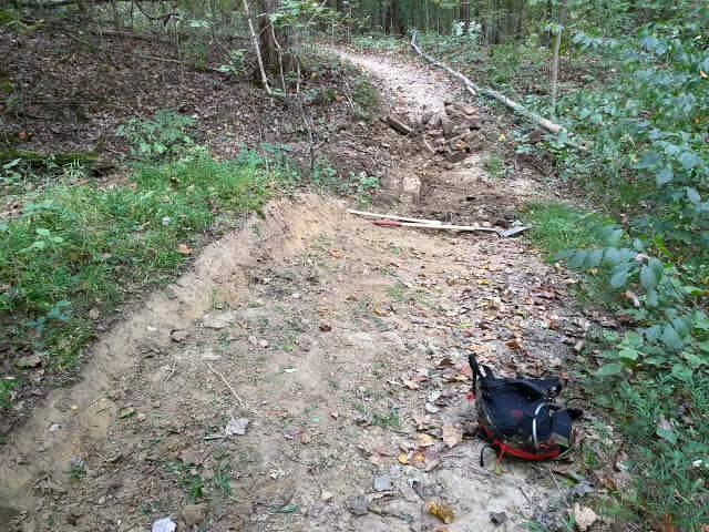 A dirt path winding through a forested area, featuring patches of grass and scattered leaves along the edges. A small pile of rocks is visible on the trail, alongside a shovel and a backpack resting on the ground. O'bannon Woods mountain bike trail.