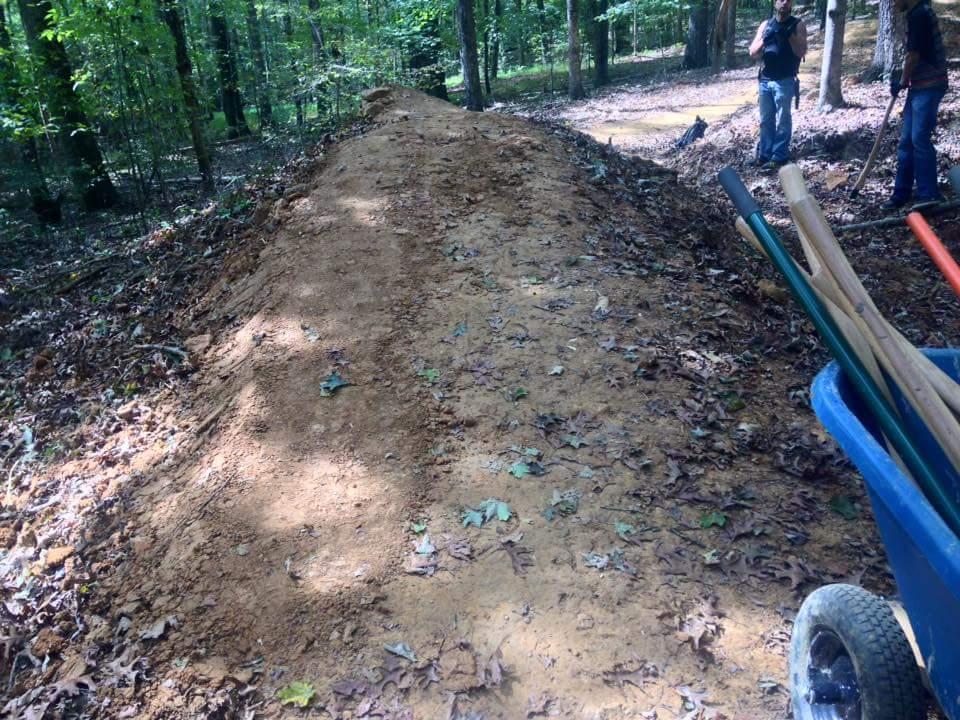 A dirt mound is visible in a wooded area, surrounded by trees and fallen leaves. In the foreground, a wheelbarrow filled with gardening tools is present, while two people can be seen in the background, engaged in outdoor work. O'bannon Woods mountain bike trail.