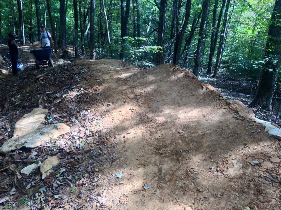 A dirt ramp is under construction in a wooded area, with two people nearby: one standing with a wheelbarrow and the other observing. Surrounding trees and fallen leaves create a natural setting. O'bannon Woods mountain bike trail.