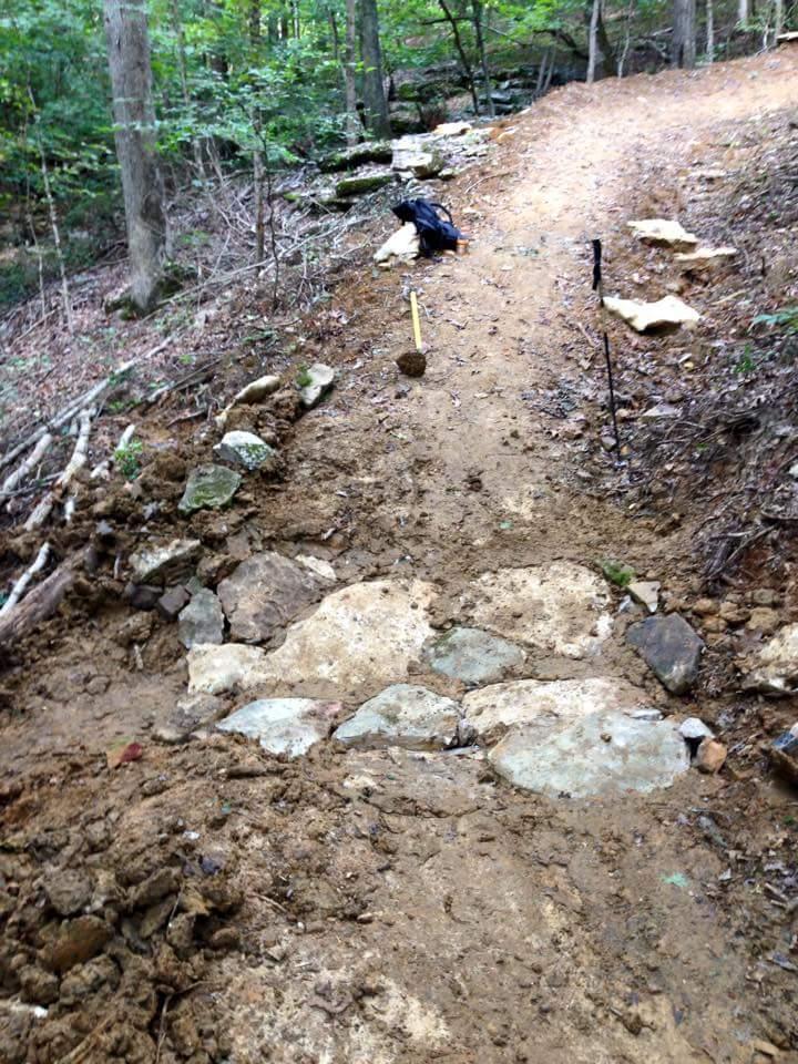 A partially cleared hiking trail with exposed rocks and dirt in a wooded area, showing ongoing trail maintenance. A black backpack and tools are visible off to the side. O'bannon Woods mountain bike trail.
