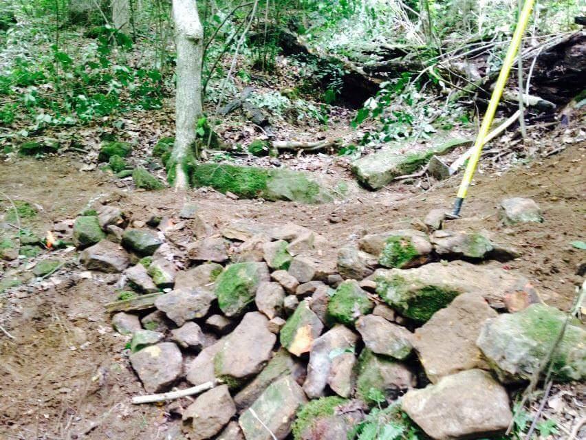 A rocky area in a forest, with a pile of moss-covered stones and dirt visible. Surrounding vegetation includes trees and leafy underbrush, indicating a natural setting. A measuring stick is partially visible on the right side of the image. O'bannon Woods mountain bike trail.