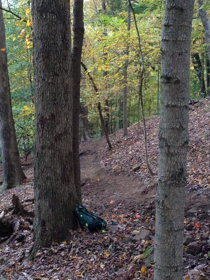 A narrow trail winds through a forest, flanked by large trees with textured bark. Fallen leaves cover the ground, and a green backpack rests against one of the trees. In the background, hints of autumn foliage can be seen, with some leaves changing color. Small orange flags mark the path, indicating a trail or route in a natural setting. O'bannon Woods mountain bike trail.