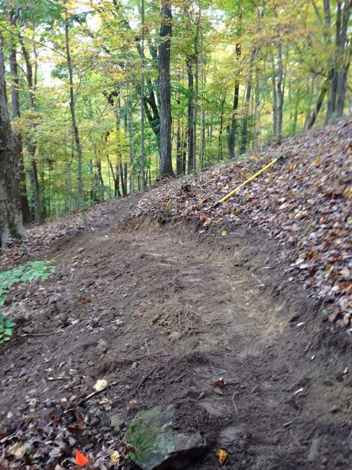 A dirt trail winding down through a wooded area, surrounded by trees with green and yellow foliage. The trail is slightly curved, showing freshly turned earth and scattered leaves. Small flags and rocks are visible along the edges. O'bannon Woods mountain bike trail.
