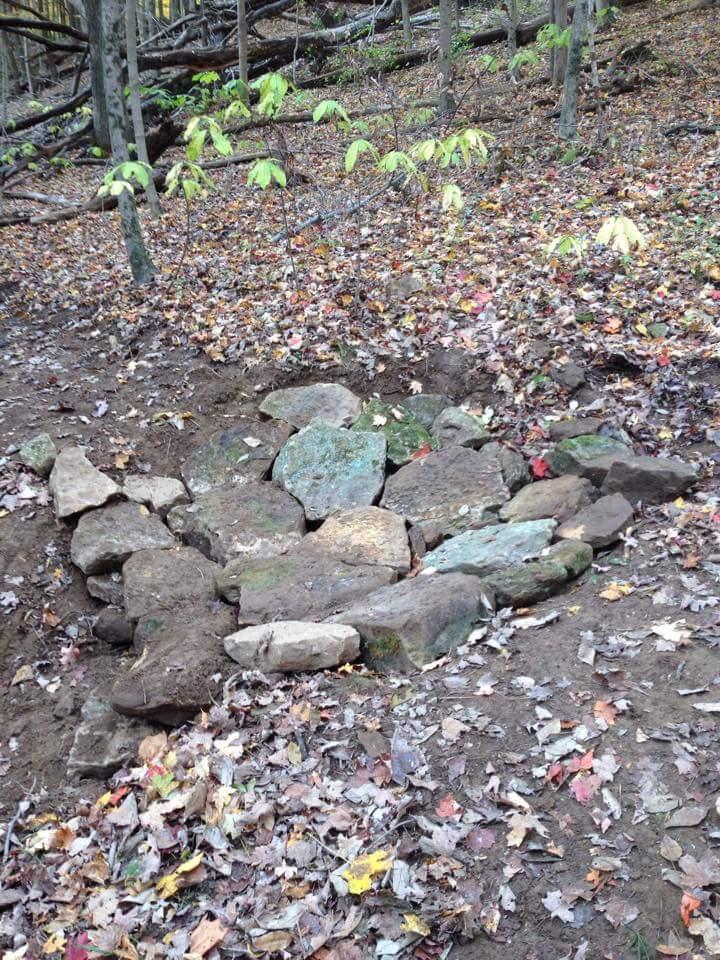 A rocky formation arranged on the ground, surrounded by fallen leaves in a forested area. The scene features trees in the background with bright green leaves, indicating early autumn. The ground is mostly bare soil mixed with different colors of fallen leaves. O'bannon Woods mountain bike trail.