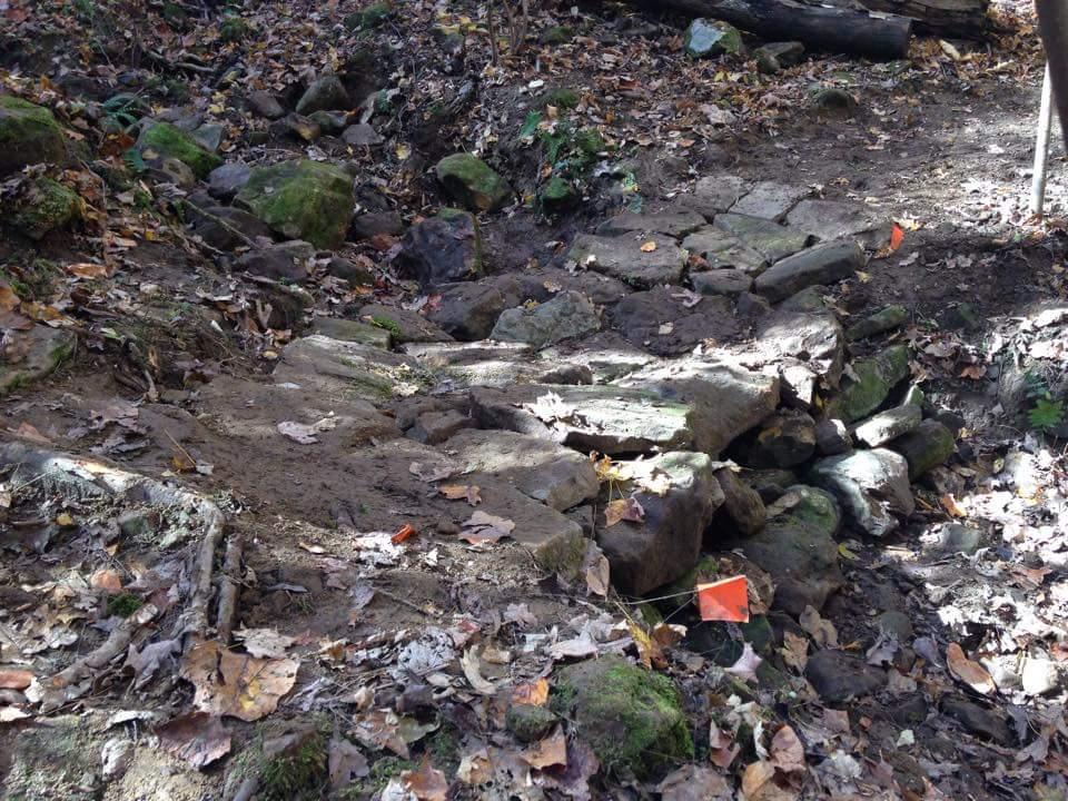 Rocky soil pathway in a wooded area, with scattered autumn leaves and small orange flags marking specific spots. O'bannon Woods mountain bike trail.