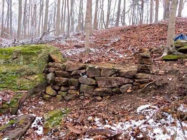 A small stone wall made of natural rocks, partially covered with moss, situated on a hillside among bare trees and fallen leaves. Snow can be seen scattered on the ground, indicating a cold, wintry environment. O'bannon Woods mountain bike trail.
