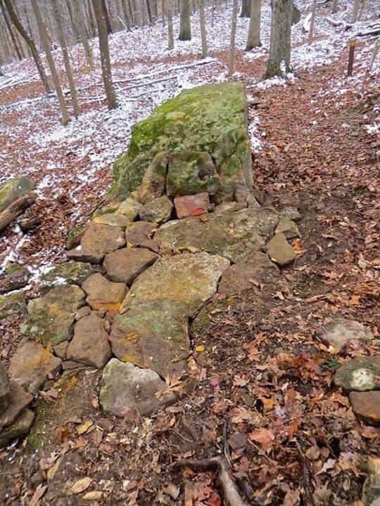Large, moss-covered rock formation surrounded by fallen leaves in a snowy forest. A path of smaller stones leads from the rock into the woodland, showcasing a blend of natural elements and terrain. O'bannon Woods mountain bike trail.