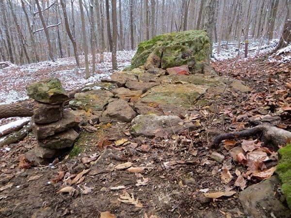 A rocky area in a forest with scattered leaves and patches of snow. A large, moss-covered boulder is prominent, accompanied by smaller stones arranged in a stack. The surrounding trees are bare, indicating a late autumn or early winter setting. O'bannon Woods mountain bike trail.