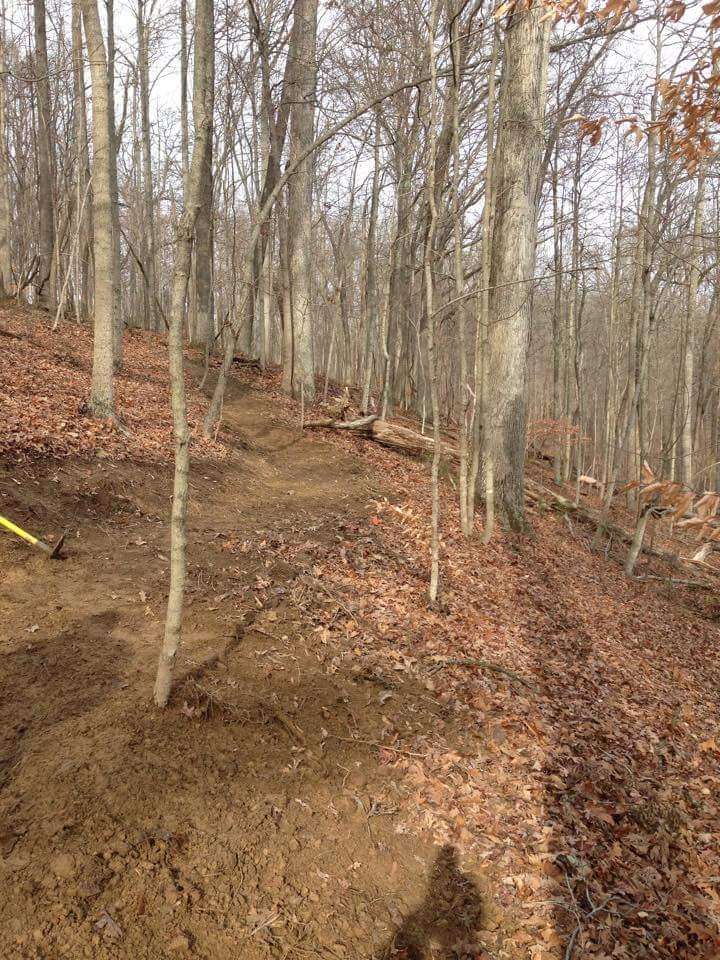 A wooded area showing a dirt path leading uphill through a landscape of bare trees and fallen leaves. A small tree is visible on the left side, and a tool, possibly a hoe or pick, is lying on the ground. The scene appears to be early spring or late fall, with a clear sky and minimal undergrowth. O'bannon Woods mountain bike trail.
