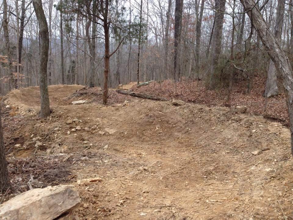 A winding dirt trail in a wooded area, with bare trees and scattered leaves on the ground. The path is surrounded by rocky terrain and natural vegetation, indicating a rustic outdoor setting. O'bannon Woods mountain bike trail.