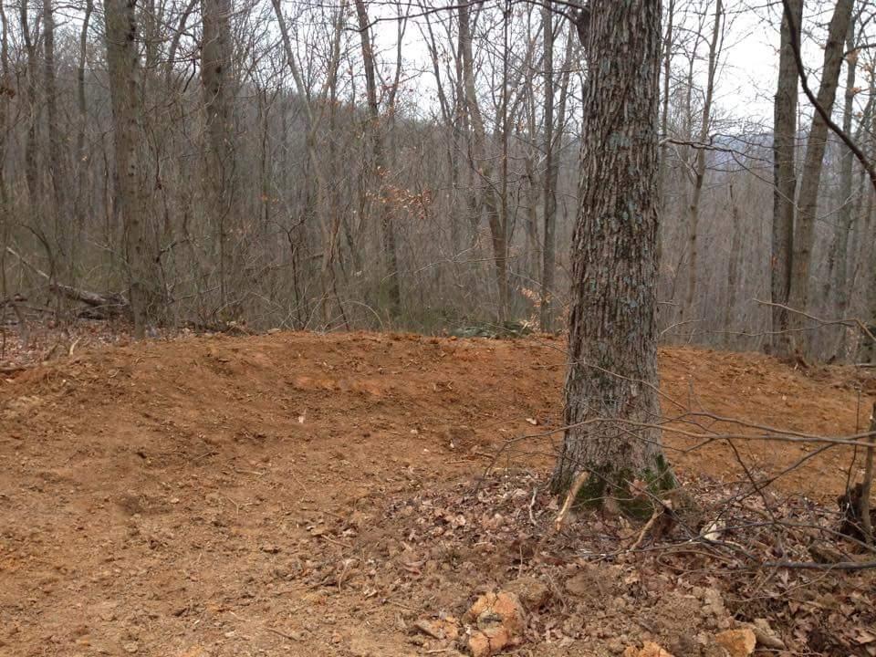 A clear area of disturbed soil surrounded by trees in a forested landscape, with some fallen leaves and twigs scattered on the ground. The scene appears to be in early spring or late fall, with bare branches on the trees and a muted, overcast sky in the background. O'bannon Woods mountain bike trail.