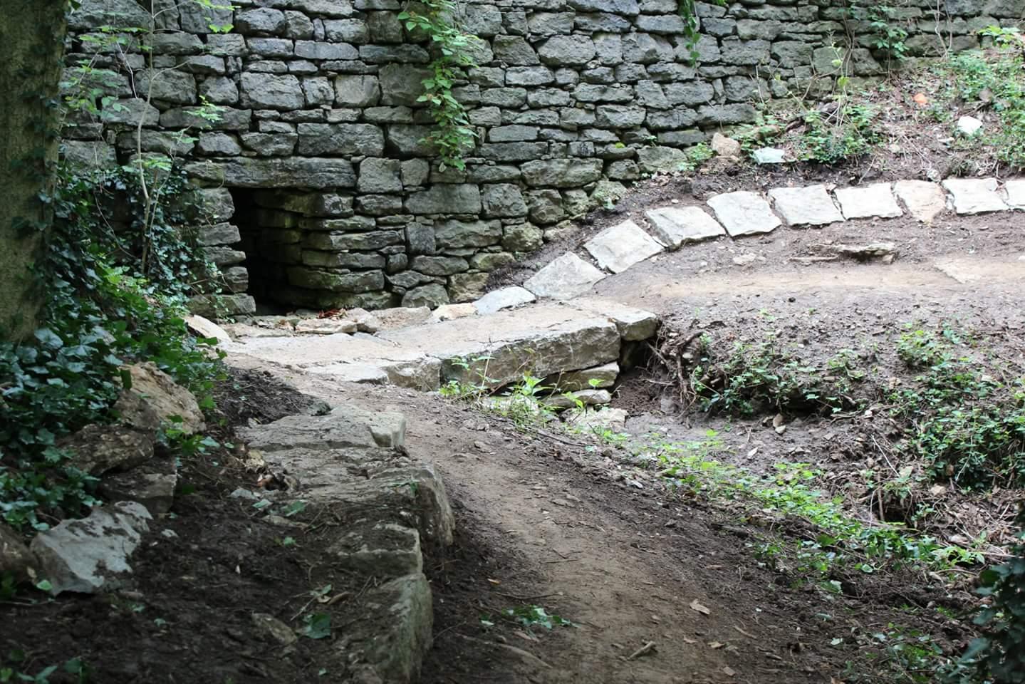 A stone wall partially covered in greenery, with a small entrance on the left. A winding path made of stones leads through a wooded area, with patches of dirt and vegetation visible along the path. Cherokee Park mountain bike trail.