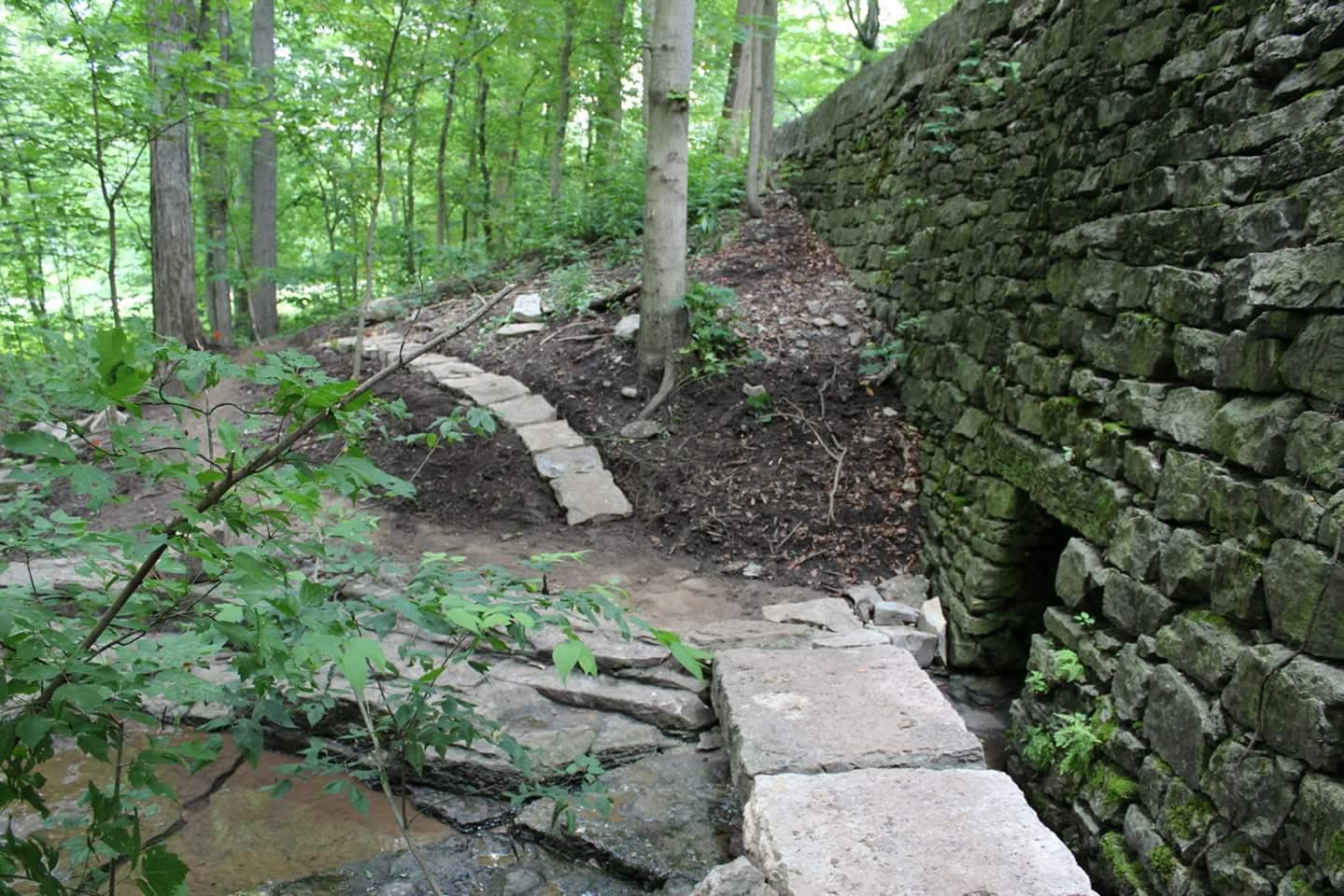 A scenic image of a forested area featuring a curved stone path leading towards a moss-covered stone wall, with greenery and trees surrounding the landscape. A small water stream is visible in the foreground, adding to the natural ambiance of the setting. Cherokee Park mountain bike trail.
