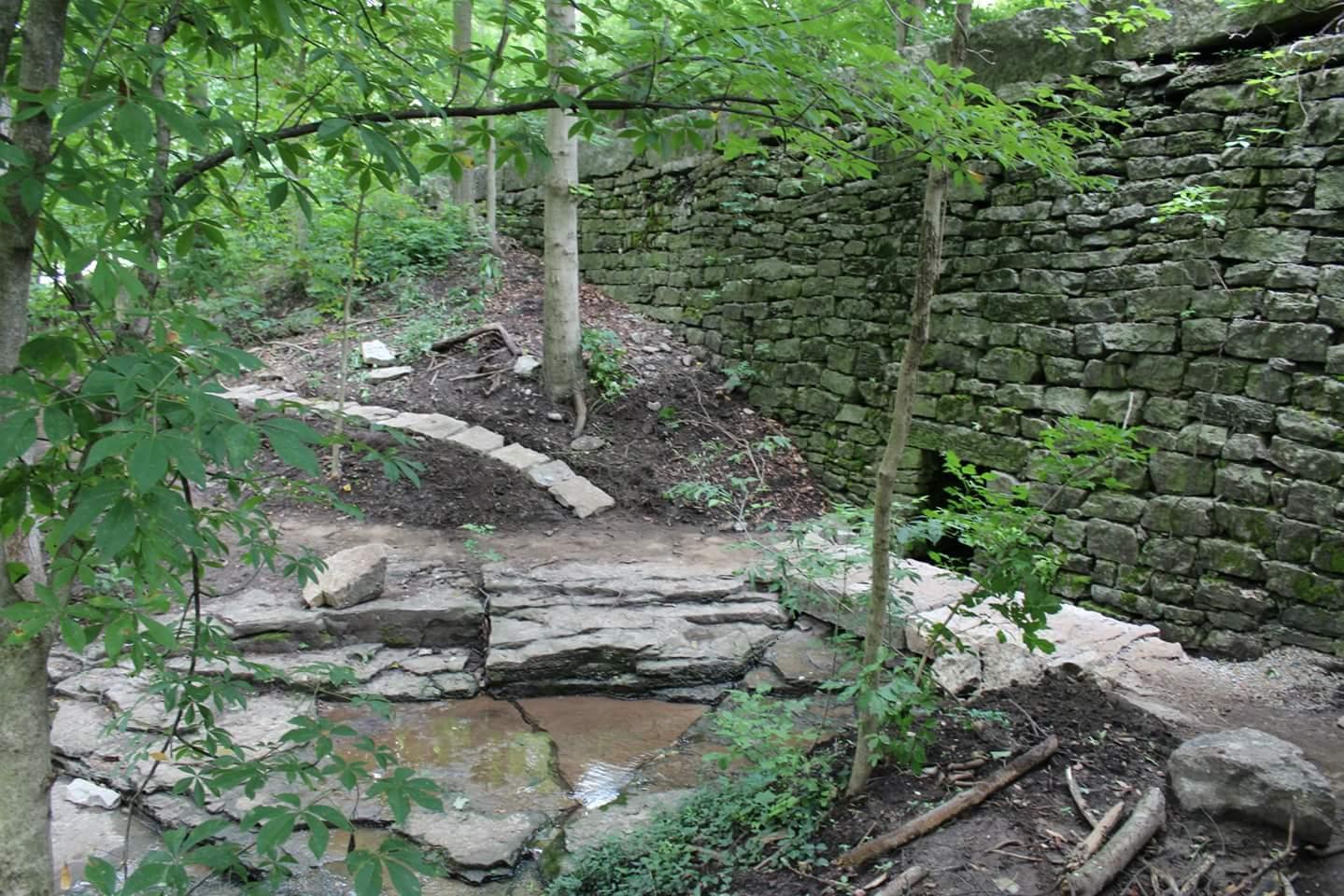 A serene wooded area featuring a curving stone pathway that leads through lush greenery. In the background, there is a weathered stone wall partially covered in moss, bordered by trees. Small rocks and patches of water can be seen along the path, creating a natural, tranquil setting. Cherokee Park mountain bike trail.