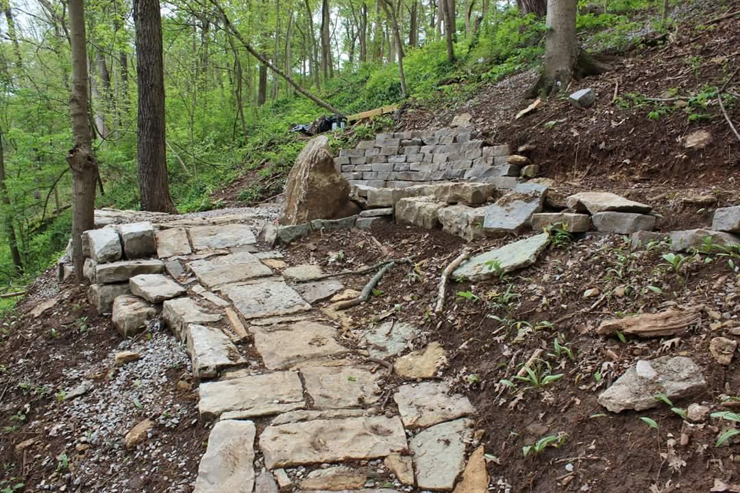 Stone path leading through a wooded area, with carefully arranged stones forming steps and a flat walkway. Lush green foliage surrounds the path, and a large boulder is visible on the side. Cherokee Park mountain bike trail.