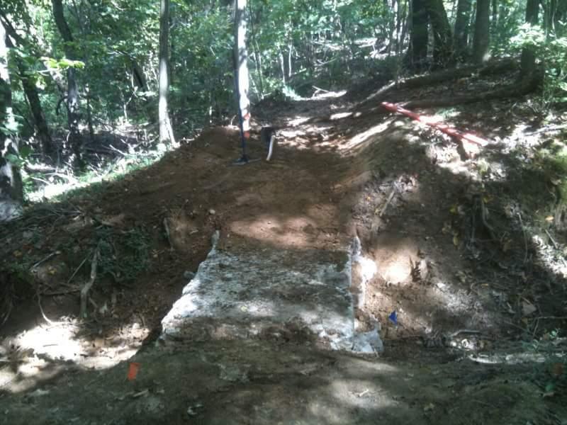 A partially cleared trail in a forested area, featuring a dirt pathway edged with dug earth, and a flat, stone-like surface at the forefront. Tools, including a shovel, are visible, suggesting ongoing construction or excavation work. Surrounding trees provide a natural backdrop. Cherokee Park mountain bike trail.