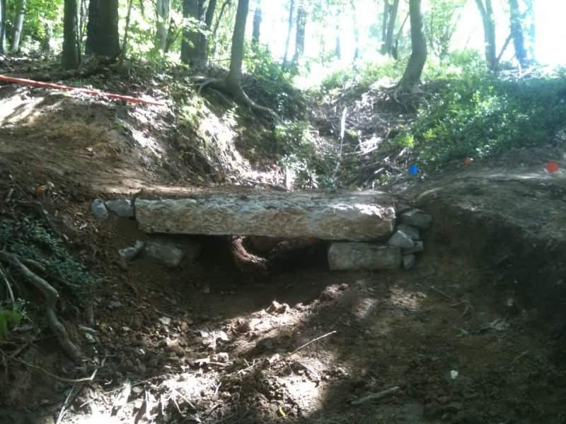 A log bridge supported by stones spans a small excavation site in a wooded area, with trees and greenery surrounding the location. Orange and blue flags mark the terrain. Cherokee Park mountain bike trail.