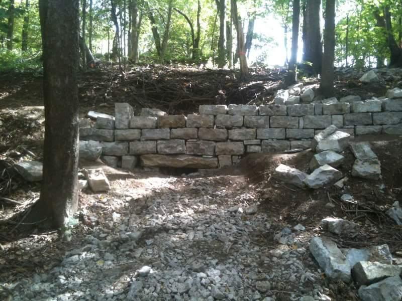 A stone wall surrounded by trees in a wooded area, with rocks scattered along a dirt path leading up to the wall. Sunlight filters through the trees, illuminating the scene. Cherokee Park mountain bike trail.