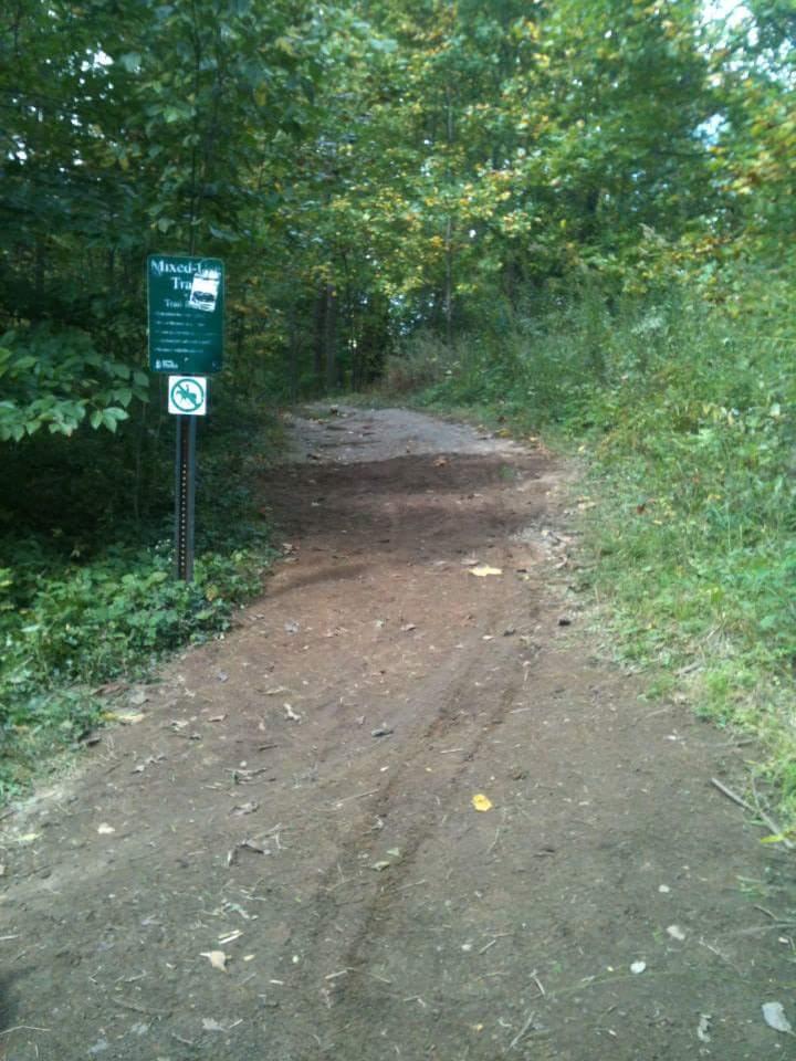 A dirt trail leading into a wooded area, marked by a sign for the "Mixed-Land Trail." The path is surrounded by lush greenery and scattered fallen leaves, indicating a natural setting. A no smoking sign is visible near the trailhead. Cherokee Park mountain bike trail.