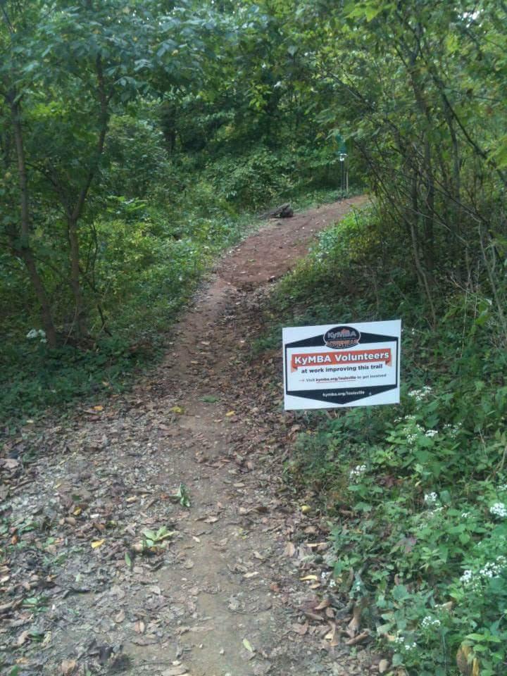 A dirt trail winding through a lush, green forest with a sign that reads "KyMBA Volunteers at work improving this trail." The path shows signs of recent maintenance. Surrounding vegetation includes trees and small plants, with a few white wildflowers visible along the edges. Cherokee Park mountain bike trail.