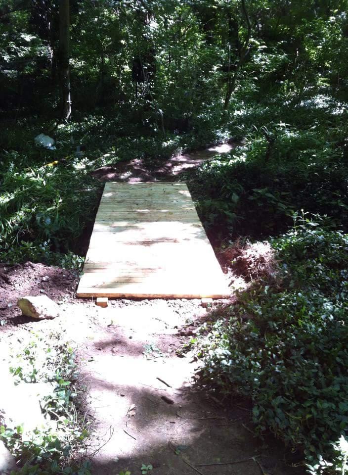 A wooden plank bridge set along a dirt path in a dense wooded area, surrounded by greenery and sunlight filtering through the trees. Cherokee Park mountain bike trail.
