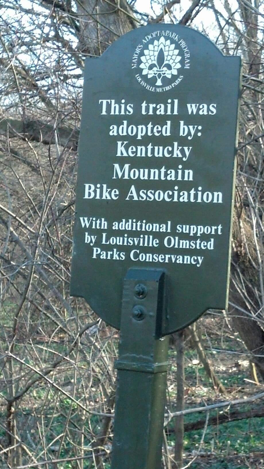 Sign indicating that a trail has been adopted by the Kentucky Mountain Bike Association, with additional support from the Louisville Olmsted Parks Conservancy. The sign is green and features a logo at the top. It is surrounded by trees and natural vegetation in a park setting. Cherokee Park mountain bike trail.