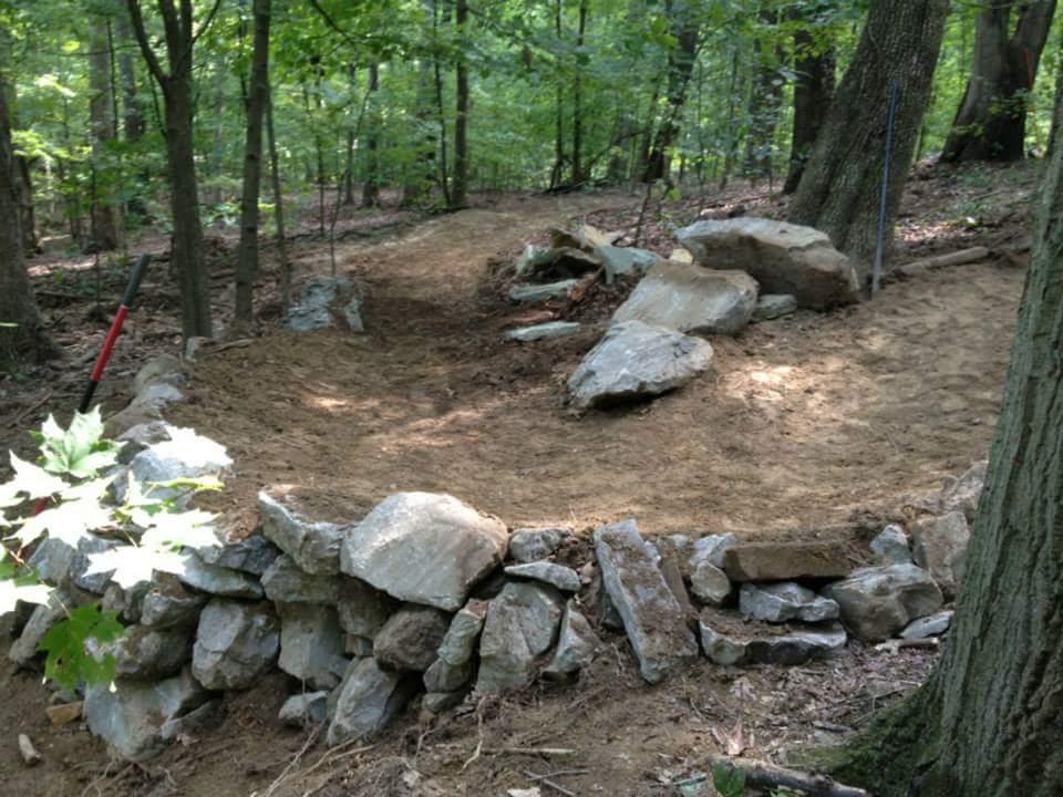 A winding dirt path in a wooded area, bordered by a stone wall and large rocks. The scene is surrounded by lush green trees, with a shovel leaning against a rock on the left side. The ground appears freshly graded and cleared, indicating recent landscaping or trail work. Waverly Park mountain bike trail.