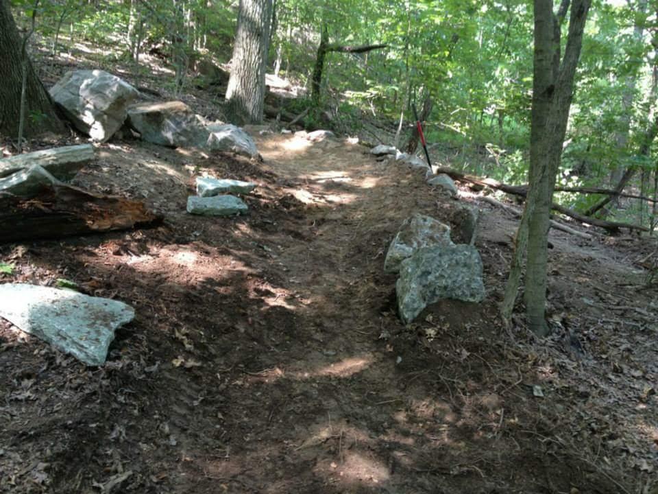 A narrow, dirt pathway meanders through a wooded area, flanked by large rocks and trees. Sunlight filters through the leaves, illuminating the trail and surrounding greenery. A red flag and tools are visible, indicating recent maintenance activity on the path. Waverly Park mountain bike trail.