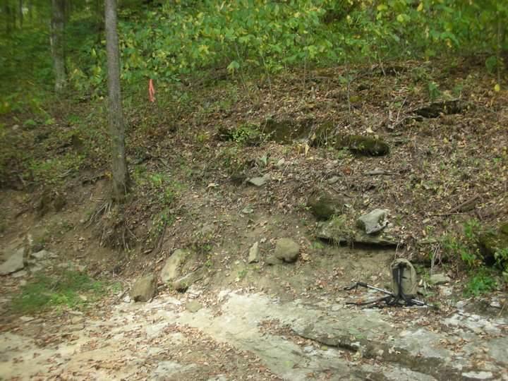 A wooded area with a sloped terrain featuring rocks and sparse vegetation. A small, abandoned backpack can be seen near the base of the slope, and a piece of bright orange tape is visible on a tree in the background. The ground is mostly bare with some fallen leaves. Brier Creek mountain bike trail.