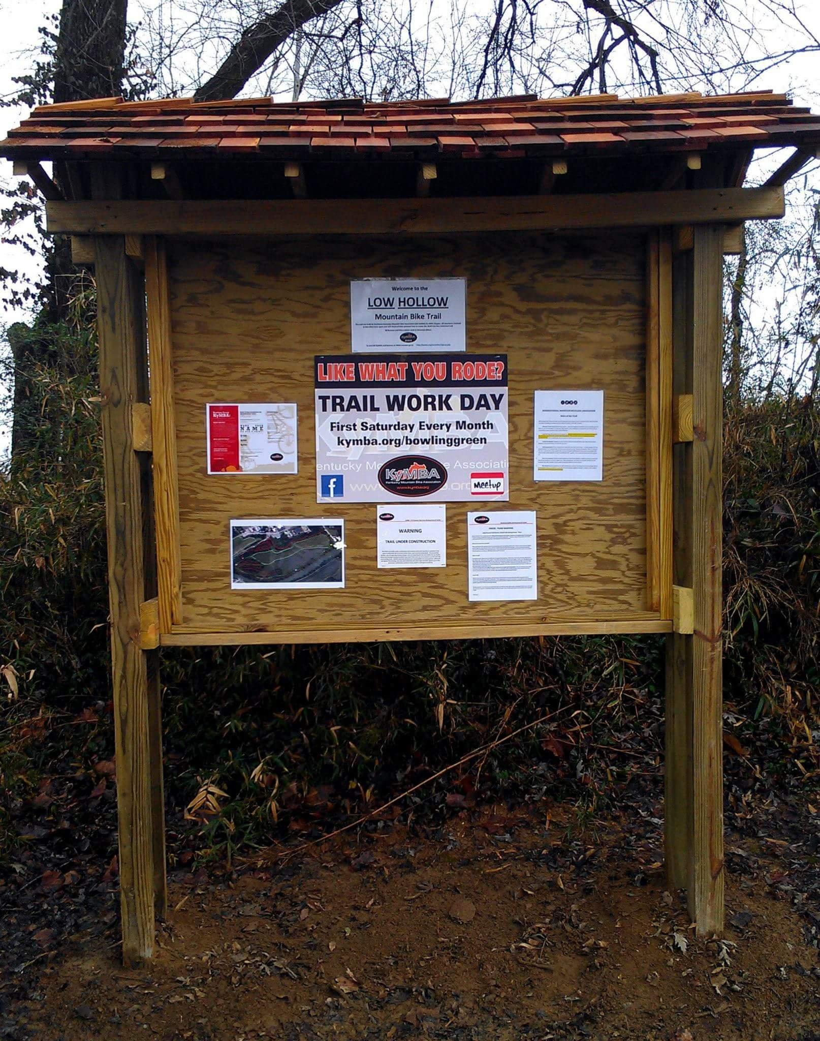 A wooden information board at the Low Hollow Mountain Bike Trail displaying announcements and event details. The sign includes a header, "LOW HOLLOW Mountain Bike Trail," and various flyers and notices about trail work days and safety warnings. The background features trees and a natural setting. Low Hollow mountain bike trail.